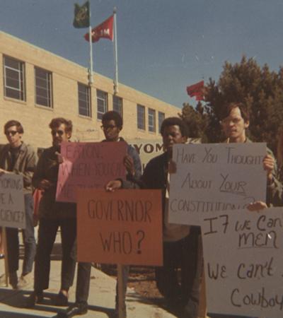 Five young male protesters, black and white, holding signs. One reads: Have you thought about your constitution?